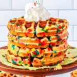 Four birthday cake waffles stacked on each other and topped with whipped cream and more sprinkles.