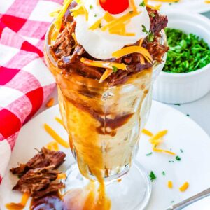 A hot beef sundae in a clear sundae dish topped with shredded chese, sour cream, and a cherry tomato.