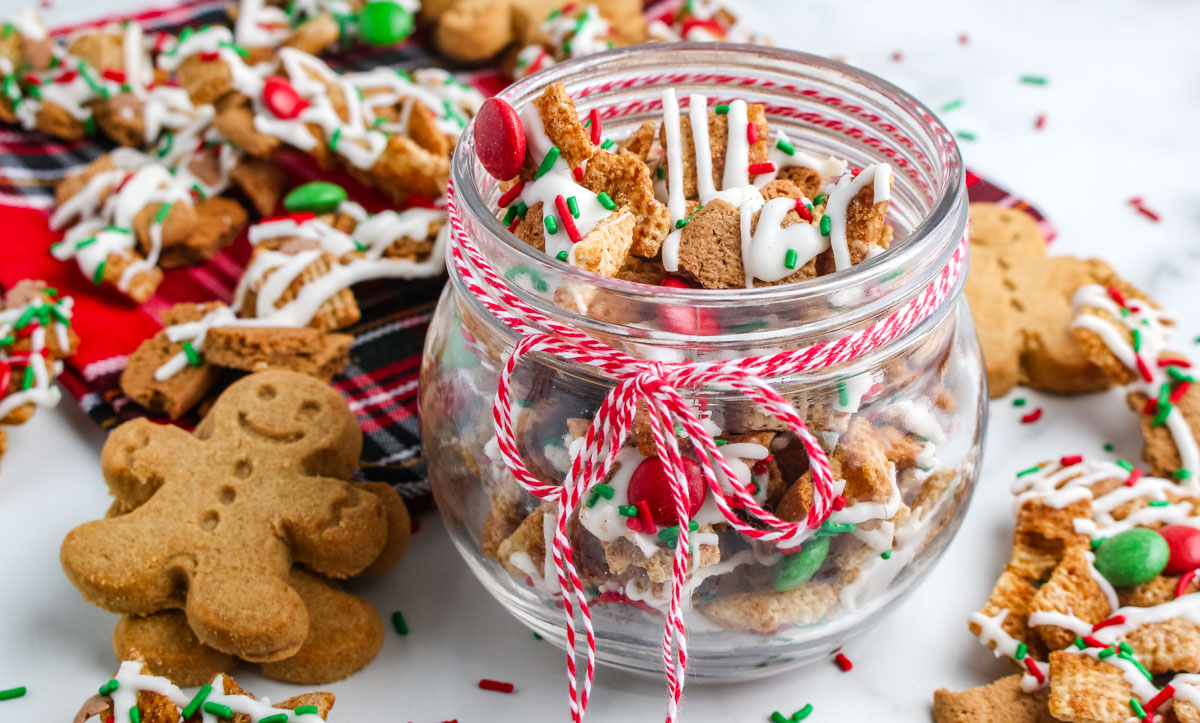 Glass jar filled with Gingerbread Chex Mix tied with red and white baker&rsquo;s twine, surrounded by gingersnap cookies and festive mix on a plaid napkin.
