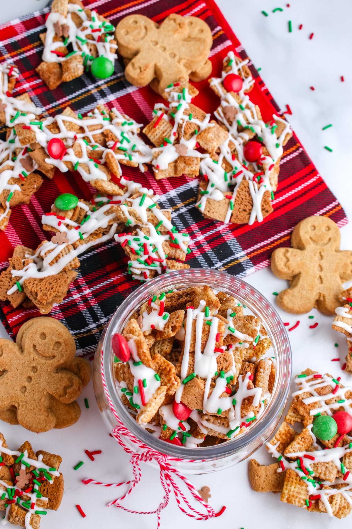 Overhead view of Gingerbread Snack Mix with Chex cereal, gingersnap pieces, red and green candies, and white chocolate drizzle scattered on a plaid cloth.