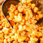A metal scoop lifting warm caramel coated puffed corn from a large mixing bowl.
