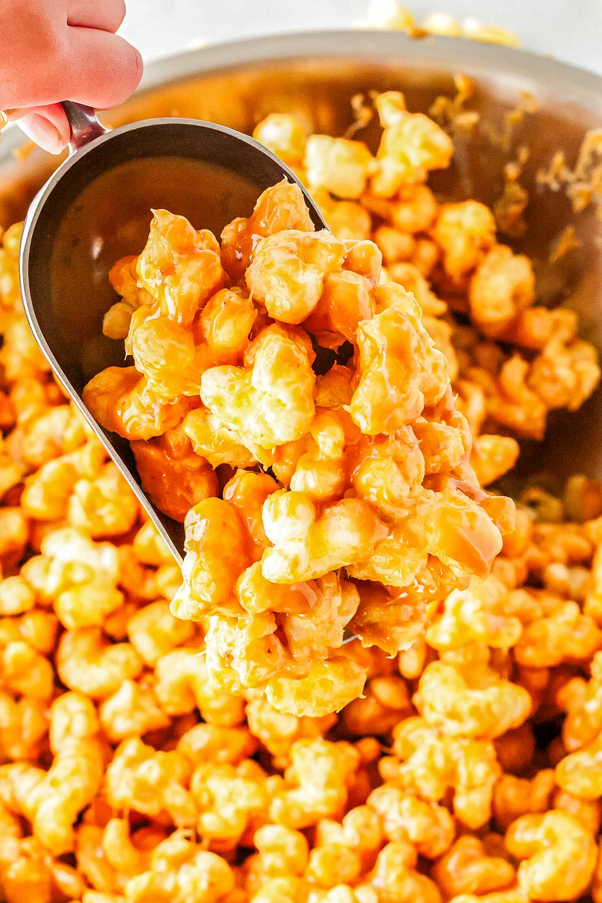 A metal scoop lifting warm caramel coated puffed corn from a large mixing bowl.