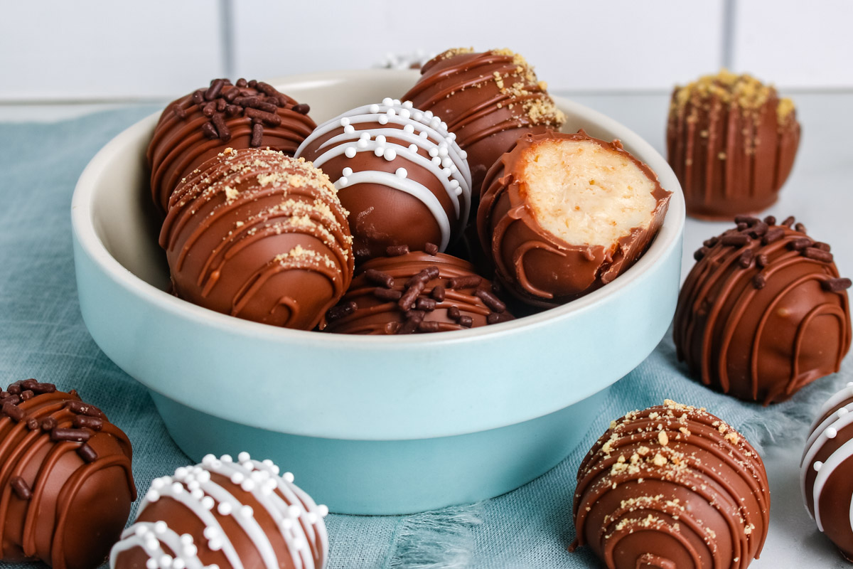 A blue bowl filled with decorated cheesecake truffles topped with sprinkles, graham crumbs, and chocolate drizzle, with extra truffles scattered on a blue napkin.