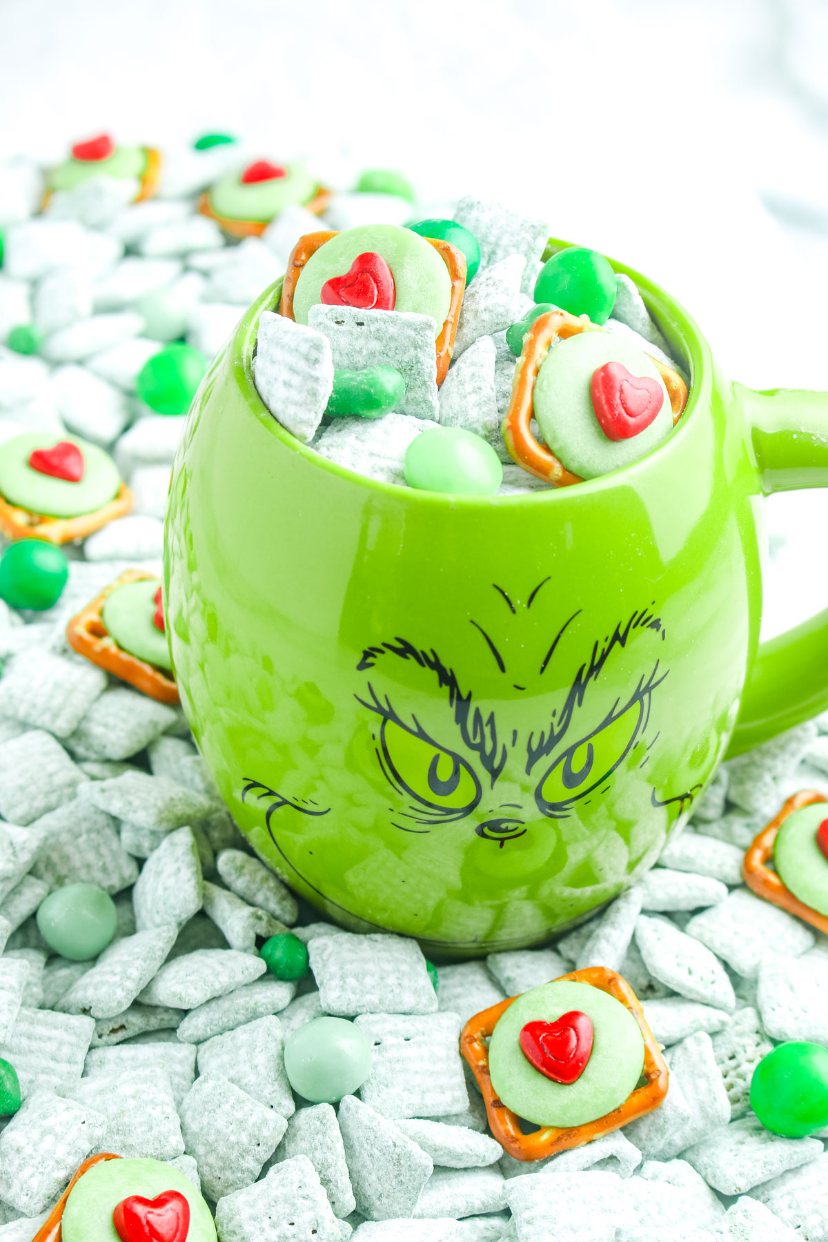 Green Grinch themed mug filled with green puppy chow, heart topped pretzels, and green candies, with more snack mix scattered across the surface.