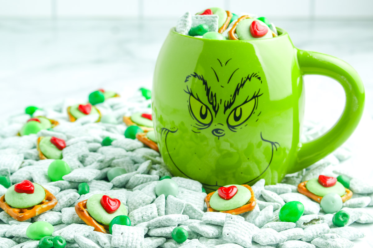 Green Grinch mug sitting on a large pile of green puppy chow, heart topped pretzels, and green candies arranged around it.