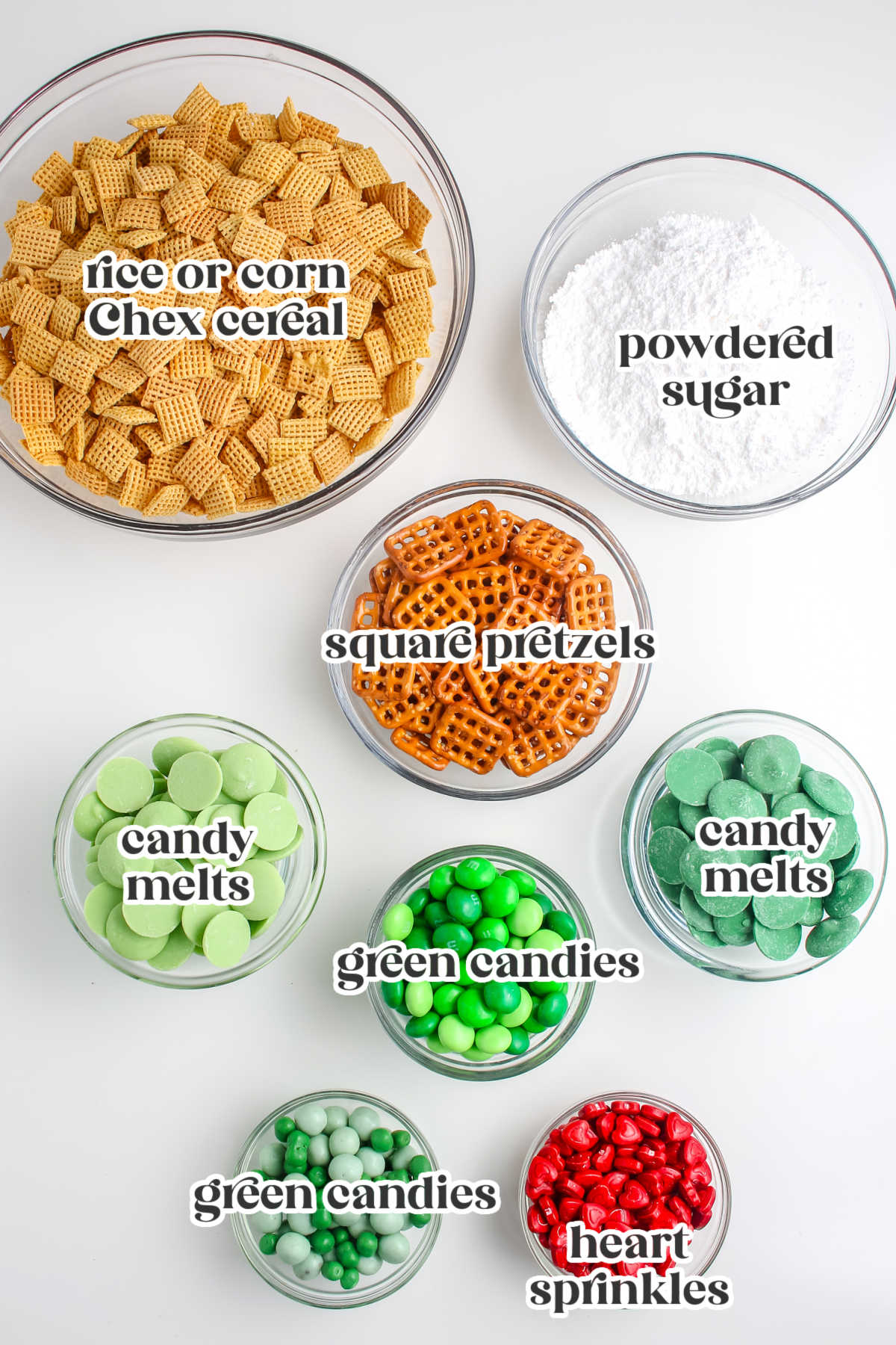Overhead display of ingredients in bowls including Chex cereal, powdered sugar, square pretzels, candy melts, heart sprinkles, and assorted green candies, each labeled for clarity.