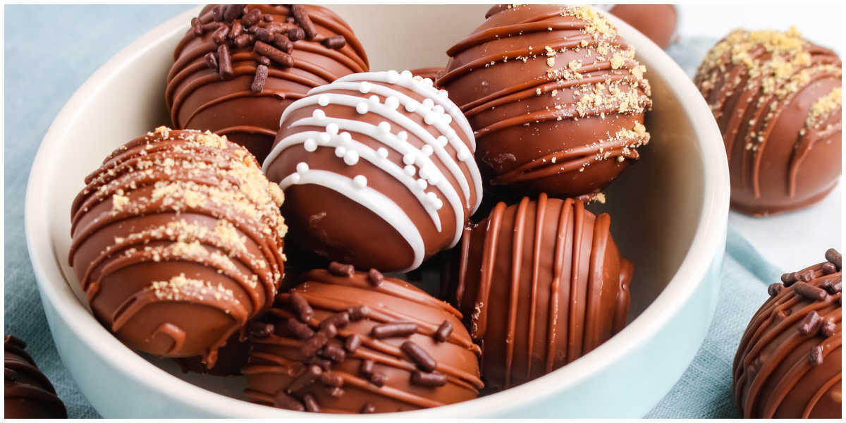 A close view of a bowl of chocolate dipped cheesecake truffles with white chocolate drizzle, sprinkles, and graham crumbs, arranged for a horizontal banner style image.