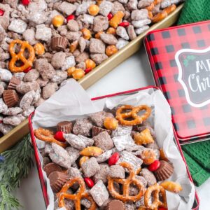 Holiday tin overflowing with Reindeer Snack Mix next to a tray of the mix, showing red candies, pretzels, puppy chow, and mini peanut butter cups.