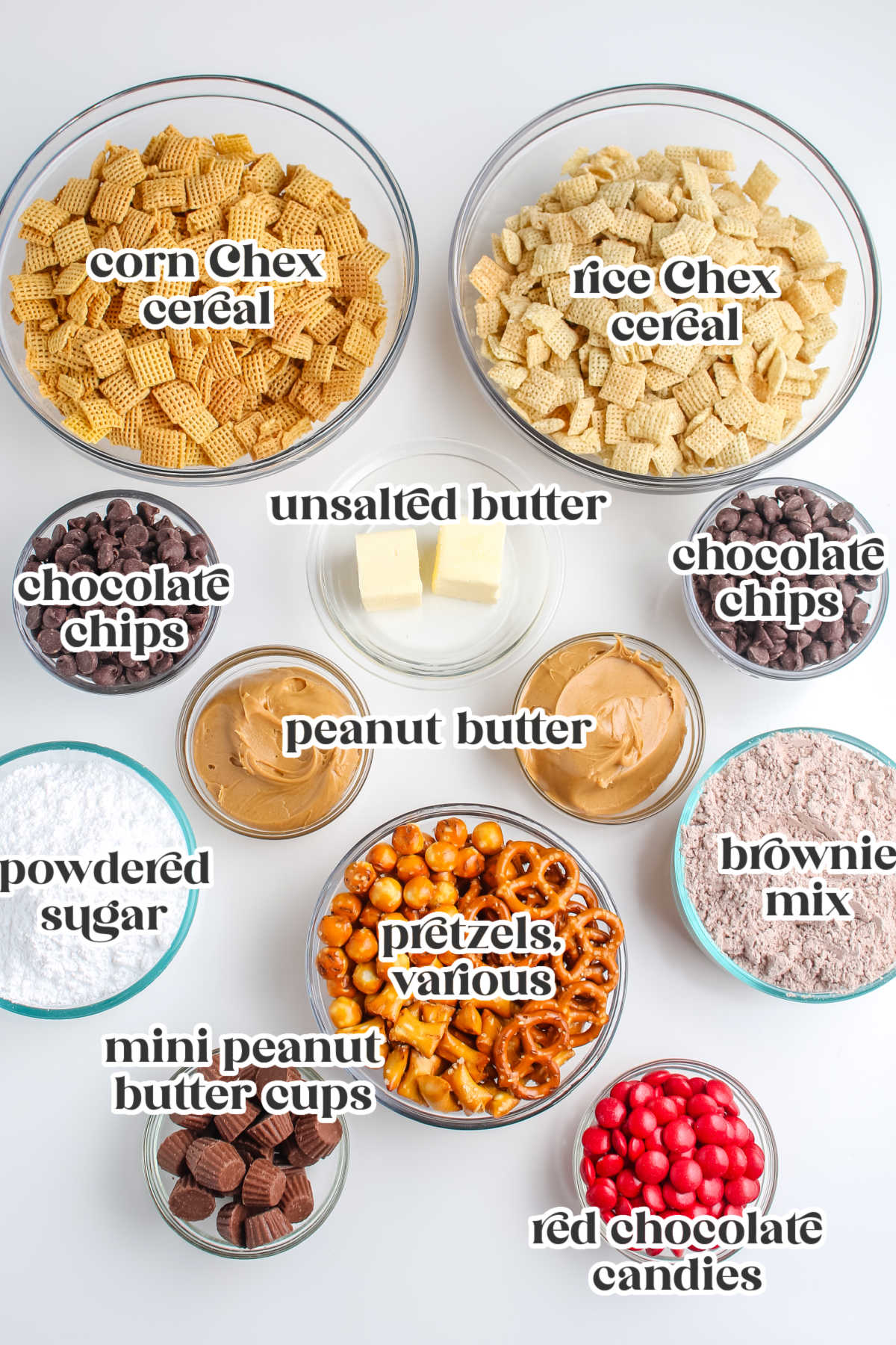 Overhead view of ingredients for Reindeer Chow, including rice and corn Chex cereal, powdered sugar, brownie mix, chocolate chips, pretzels, peanut butter, butter, mini peanut butter cups, and red chocolate candies.