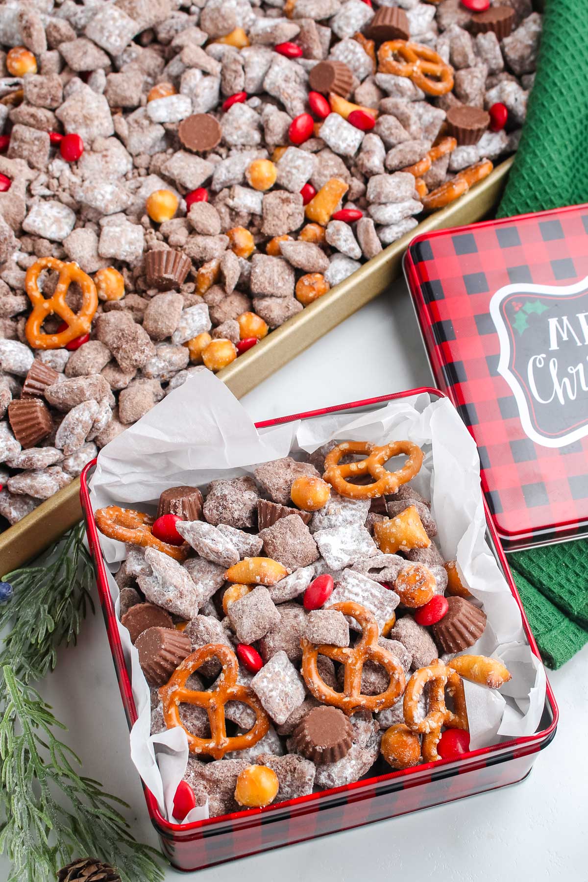 Holiday tin overflowing with Reindeer Snack Mix next to a tray of the mix, showing red candies, pretzels, puppy chow, and mini peanut butter cups.