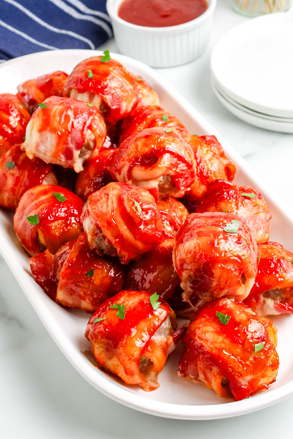 A white platter piled high with sweet and smoky bacon wrapped meatballs, glazed with a brown sugar sauce and garnished with parsley.