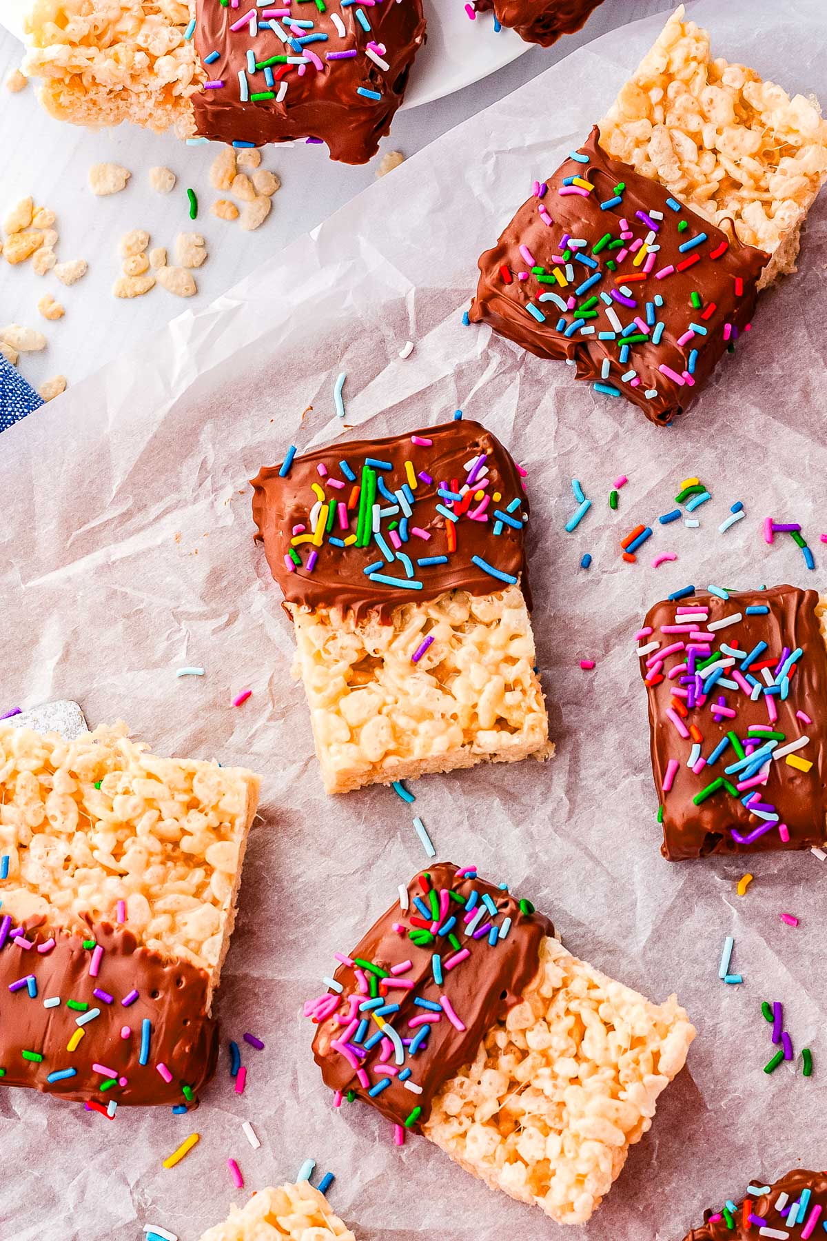 A stack of ultra-chewy Rice Krispie treats dipped halfway in melted milk chocolate and topped with festive sprinkles, served on a white dessert platter.