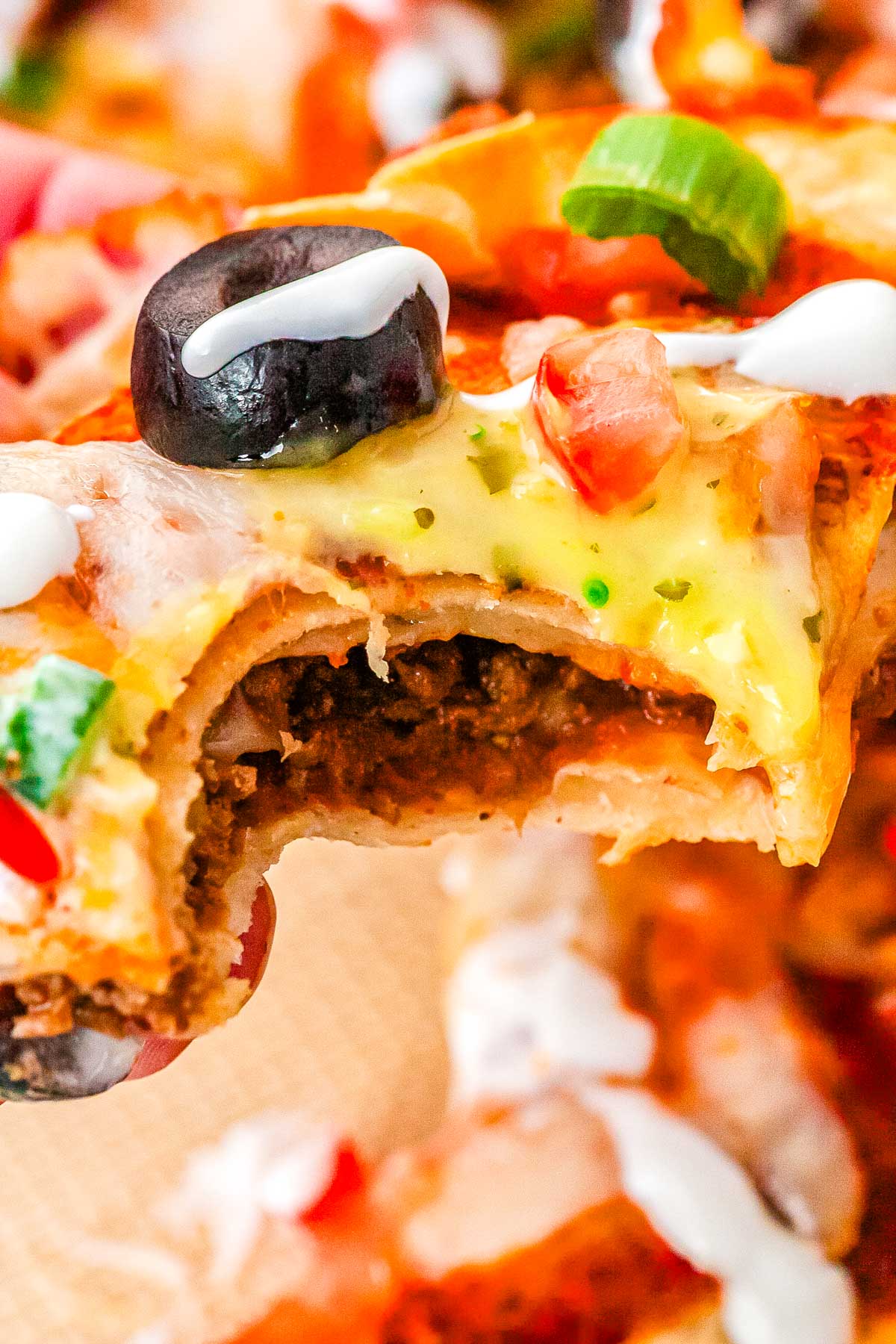Close up of a homemade Mexican Pizza slice with a bite taken out of it showing layers of crispy tortillas, seasoned beef, melted cheese, diced tomatoes, olives, green onion, and sour cream.
