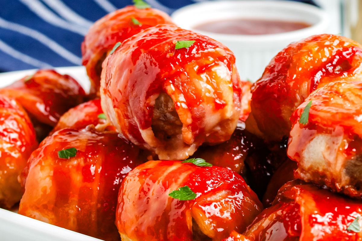 Close-up view of glazed bacon wrapped meatballs on a platter, showing the crispy bacon edges and sticky red sauce.