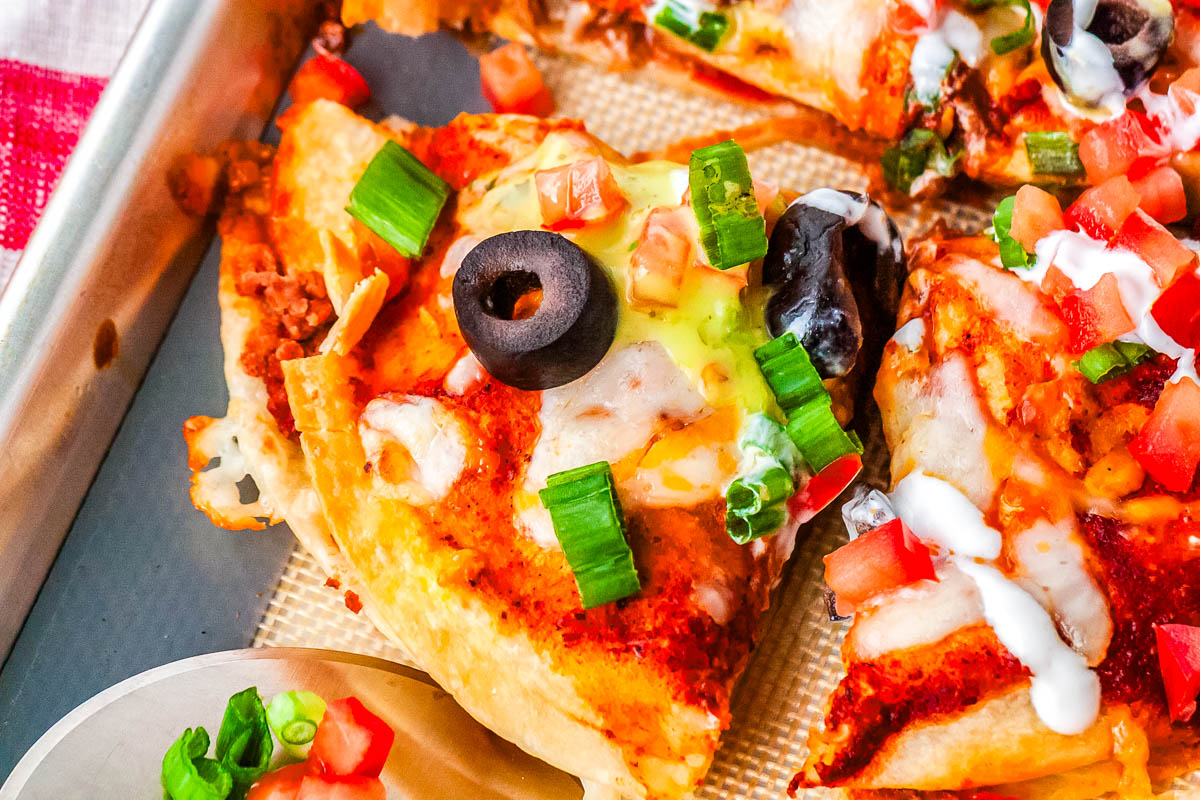 Slice of homemade Mexican Pizza topped with melted cheese, diced tomatoes, sliced olives, green onion, and sour cream on a sheet pan.