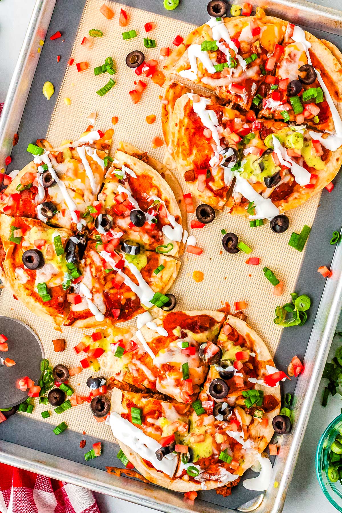 Three homemade Mexican Pizzas on a sheet pan, each sliced and topped with tomatoes, olives, green onion, sour cream, and guacamole.