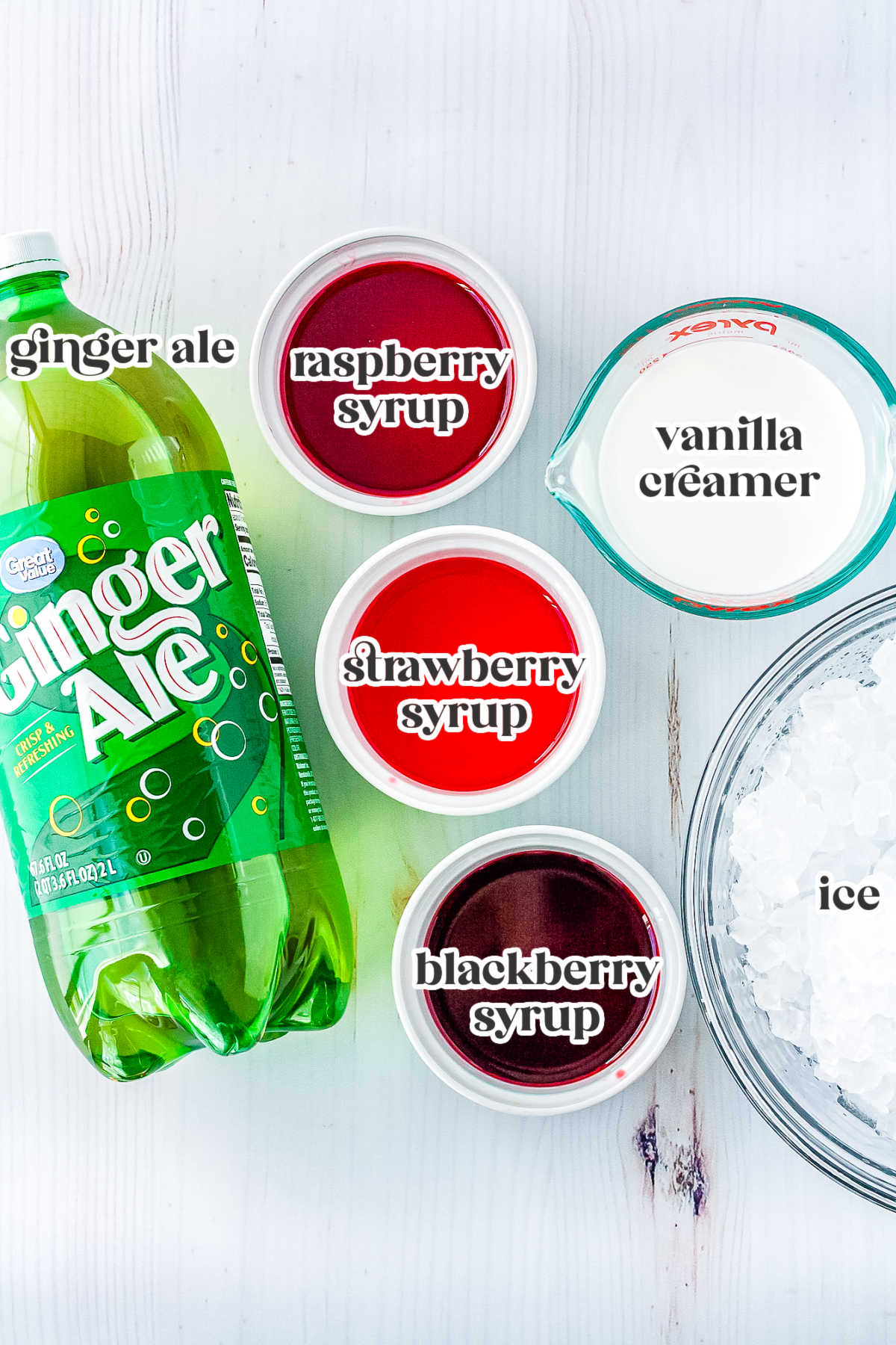 Ingredients for Polar Punch laid out on a white surface including ginger ale, berry syrups, vanilla creamer, and a bowl of ice.