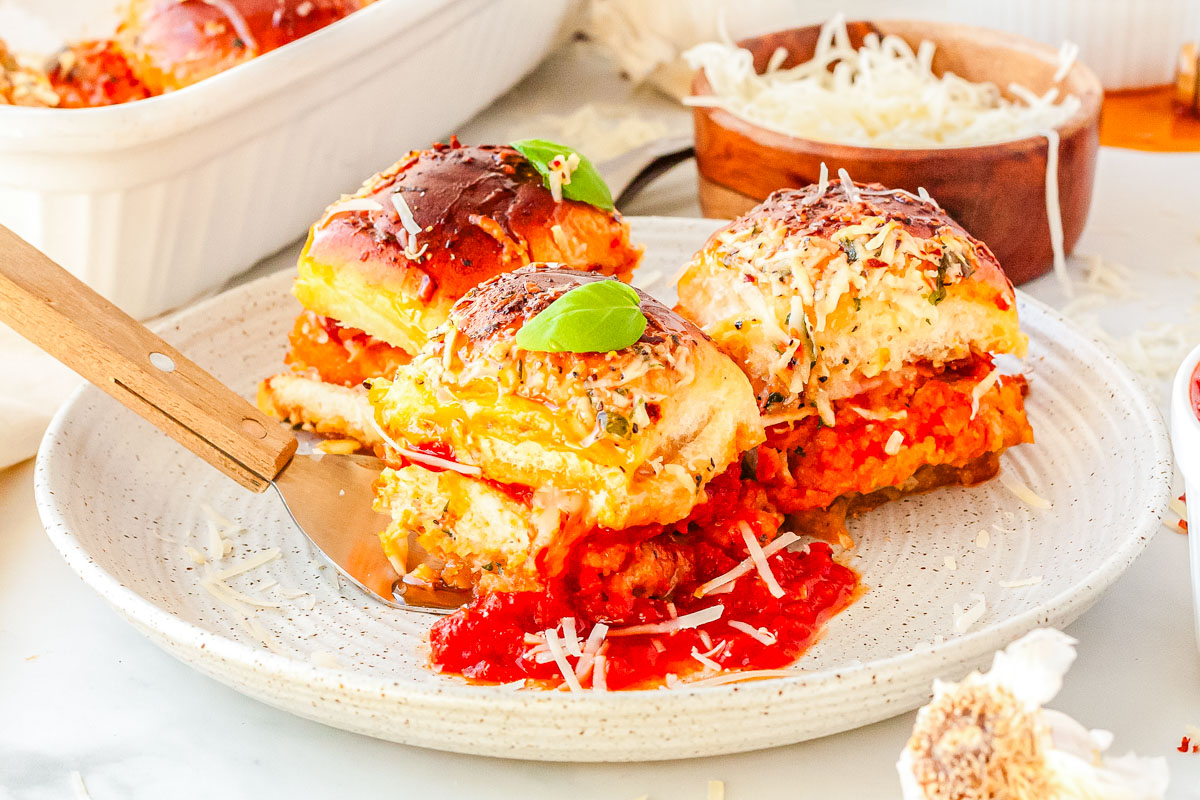 Three warm chicken parmesan sliders served on a speckled plate with a side of marinara sauce for dipping.