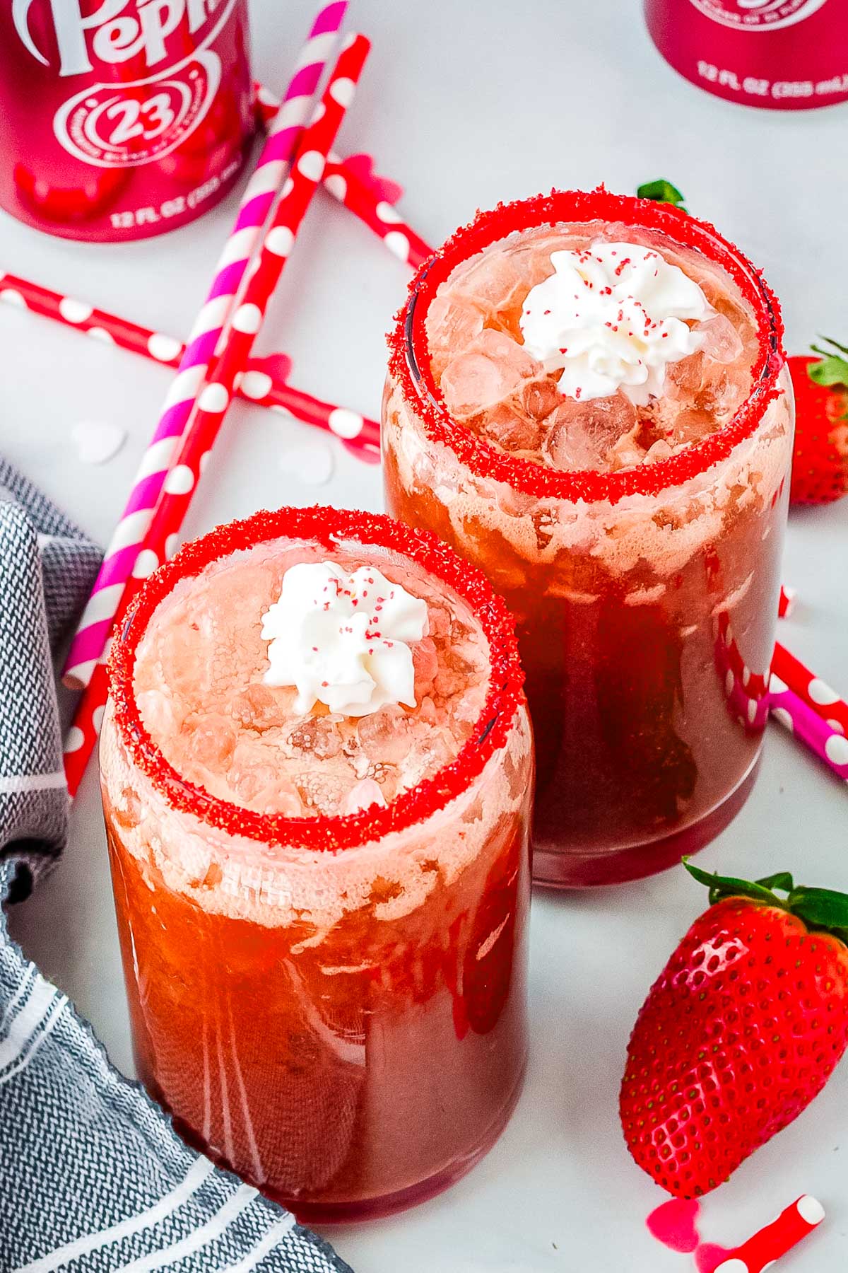 Valentine&rsquo;s Day strawberry dirty Dr Pepper drinks in clear glasses with pebble ice, red sugar rims, whipped cream, and fresh strawberries on a white surface.