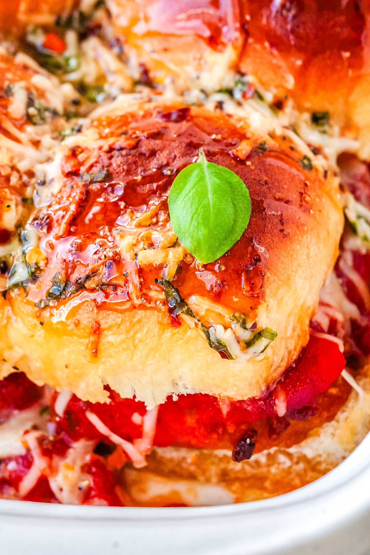 A macro shot of a golden brown slider bun topped with a glossy hot honey glaze, crushed red pepper flakes, and a fresh basil leaf.