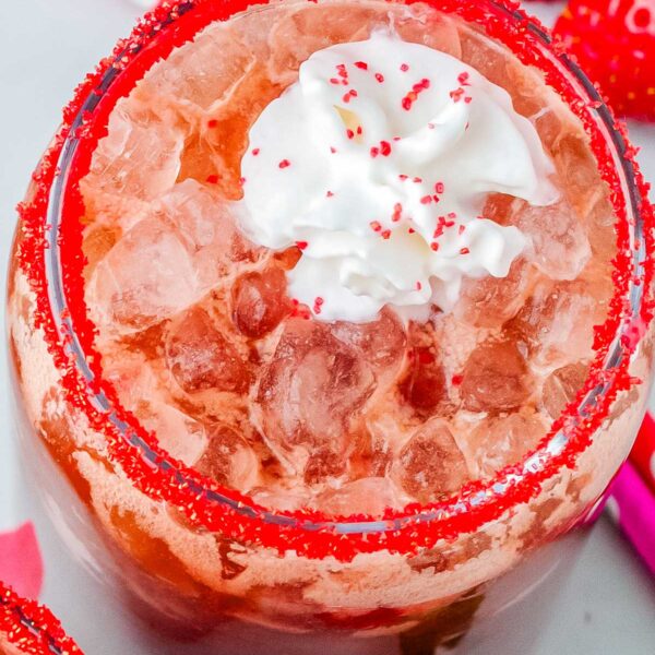 Strawberry dirty Dr Pepper drink with pebble ice and whipped cream, garnished with red sugar rim and surrounded by fresh strawberries.