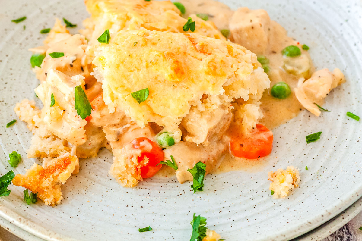 Scoop of chicken pot pie with cheddar biscuit topping on a plate, showing creamy chicken filling with peas and carrots.
