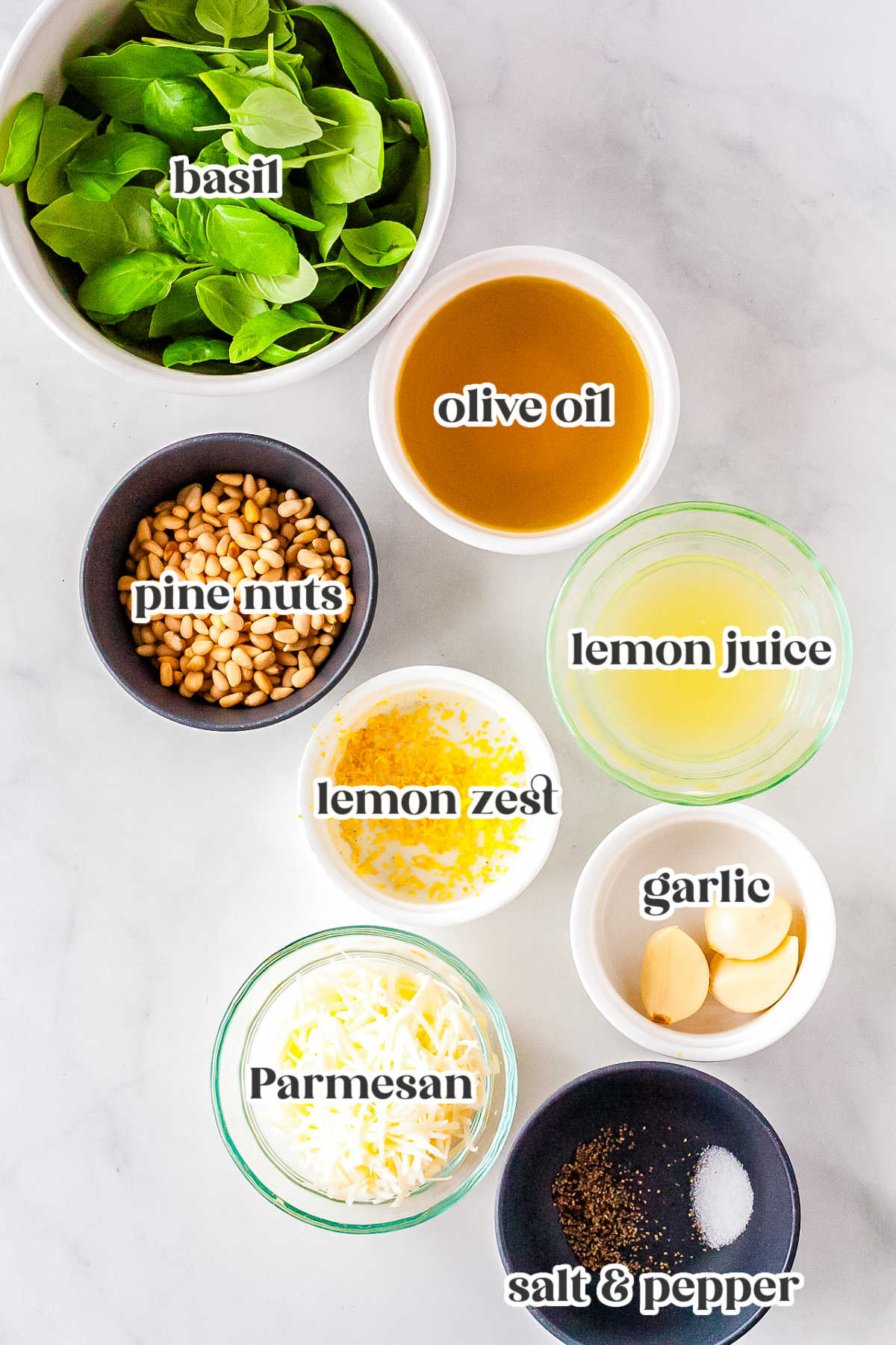 A flat lay of the ingredients needed for the recipe including fresh basil, olive oil, pine nuts, lemon juice, lemon zest, garlic cloves, Parmesan cheese, salt, and black pepper.
