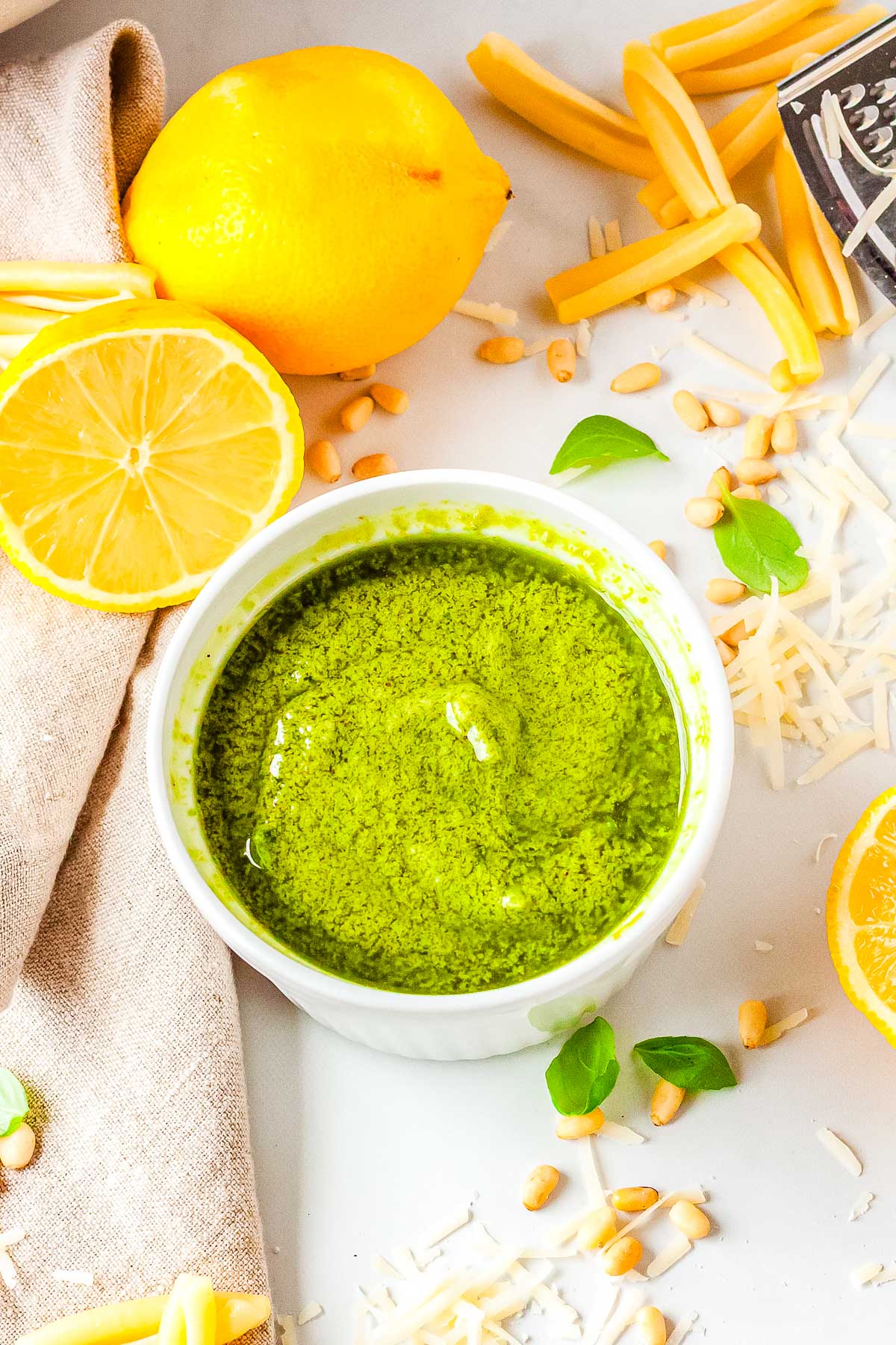 An overhead view of a white bowl filled with bright green lemon pesto sauce surrounded by fresh lemons, pine nuts, basil leaves, and grated Parmesan.