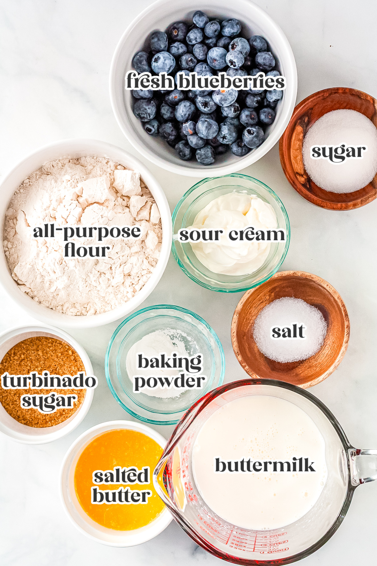 Ingredients for blueberry swim biscuits including fresh blueberries, flour, sugar, sour cream, buttermilk, melted butter, baking powder, salt, and turbinado sugar.