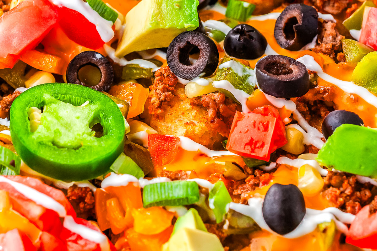 Close up of loaded totchos with melted cheese, ground beef, jalape&ntilde;os, olives, tomatoes, avocado, and sour cream drizzle.