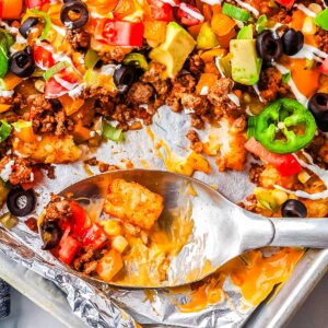 Serving spoon scooping cheesy totchos with ground beef, melted cheese, and diced vegetables from a sheet pan.