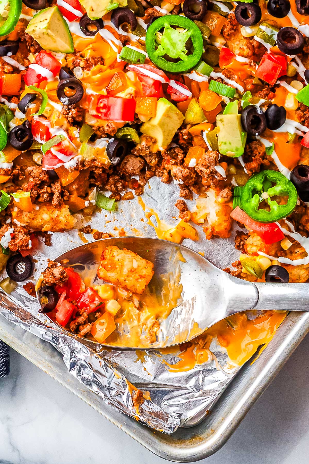 Serving spoon scooping cheesy totchos with ground beef, melted cheese, and diced vegetables from a sheet pan.