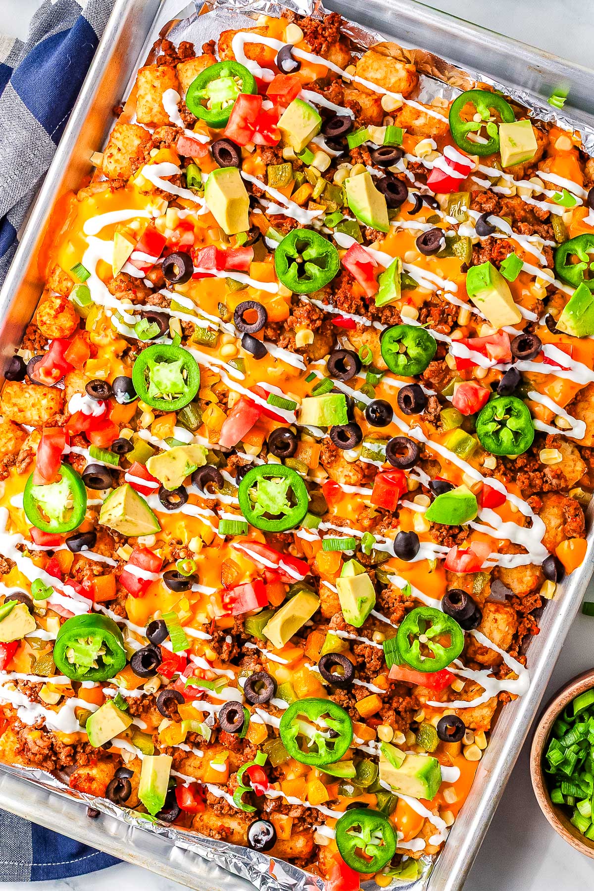 Fully loaded totchos on a sheet pan topped with jalape&ntilde;os, olives, avocado, tomatoes, green onions, and sour cream drizzle.
