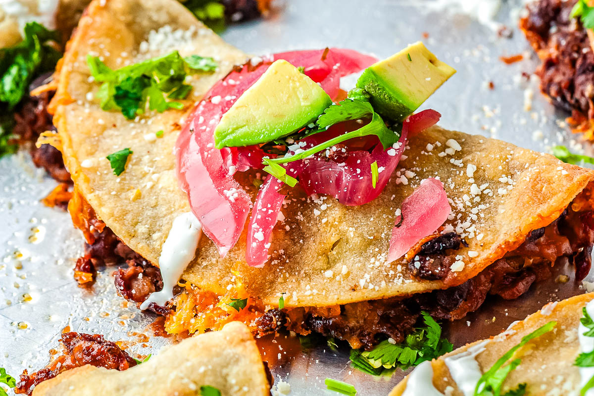 Crispy baked black bean taco topped with avocado, pickled red onions, cilantro, and cotija cheese on a sheet pan.