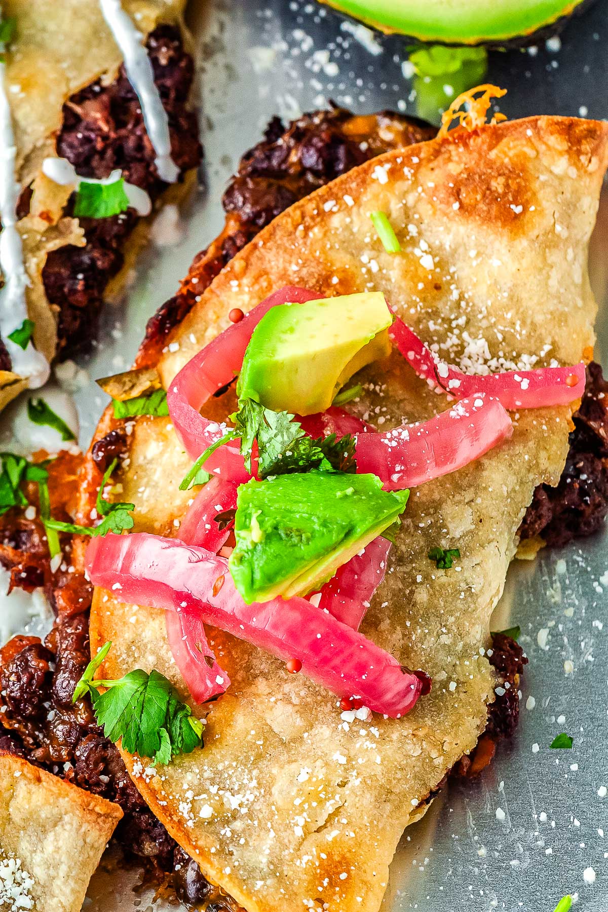 Close up of a golden crispy black bean taco filled with seasoned beans and cheddar cheese topped with avocado and pickled onions.