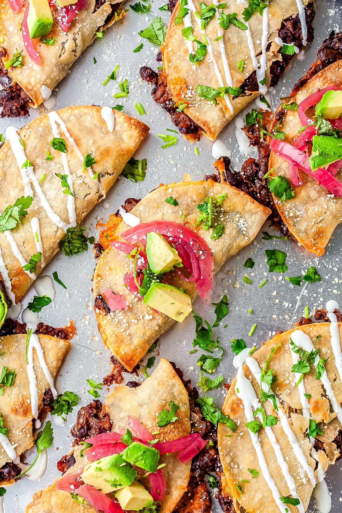 Overhead view of crispy black bean tacos on a baking sheet garnished with cilantro, avocado, pickled onions, and sour cream drizzle.
