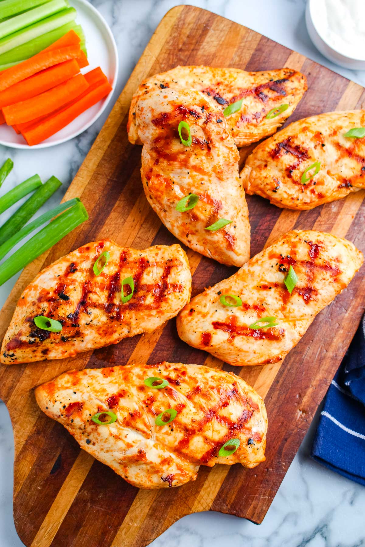 Grilled buffalo chicken breasts arranged on a wooden cutting board with green onions, carrots, celery, and ranch dressing.