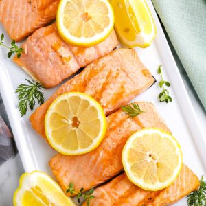 Overhead view of grilled salmon fillets garnished with lemon slices and fresh herbs on a white serving platter.