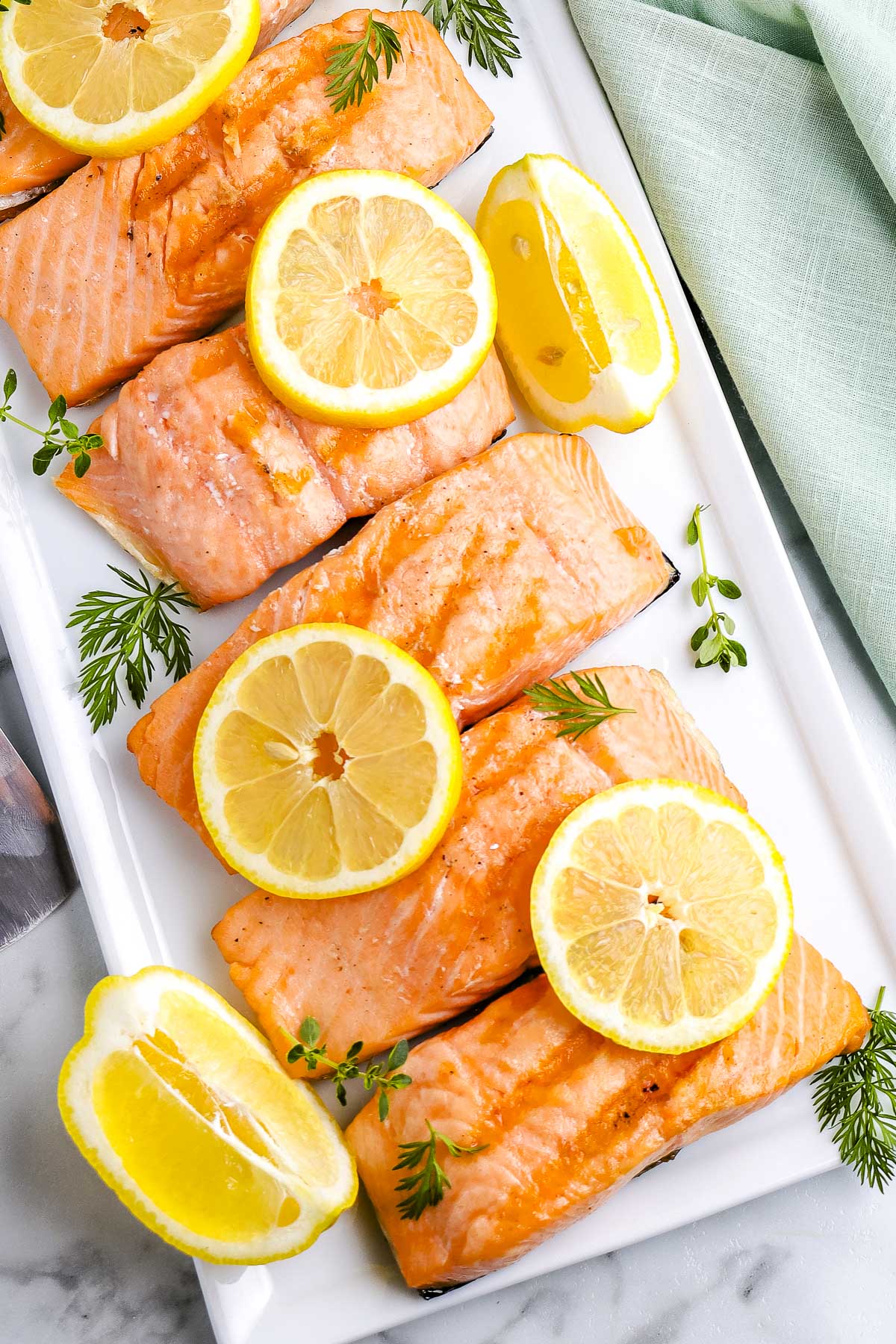 Overhead view of grilled salmon fillets garnished with lemon slices and fresh herbs on a white serving platter.