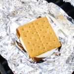 Unwrapped grilled s'more assembled with graham crackers, chocolate, and a marshmallow on aluminum foil over a grill grate.