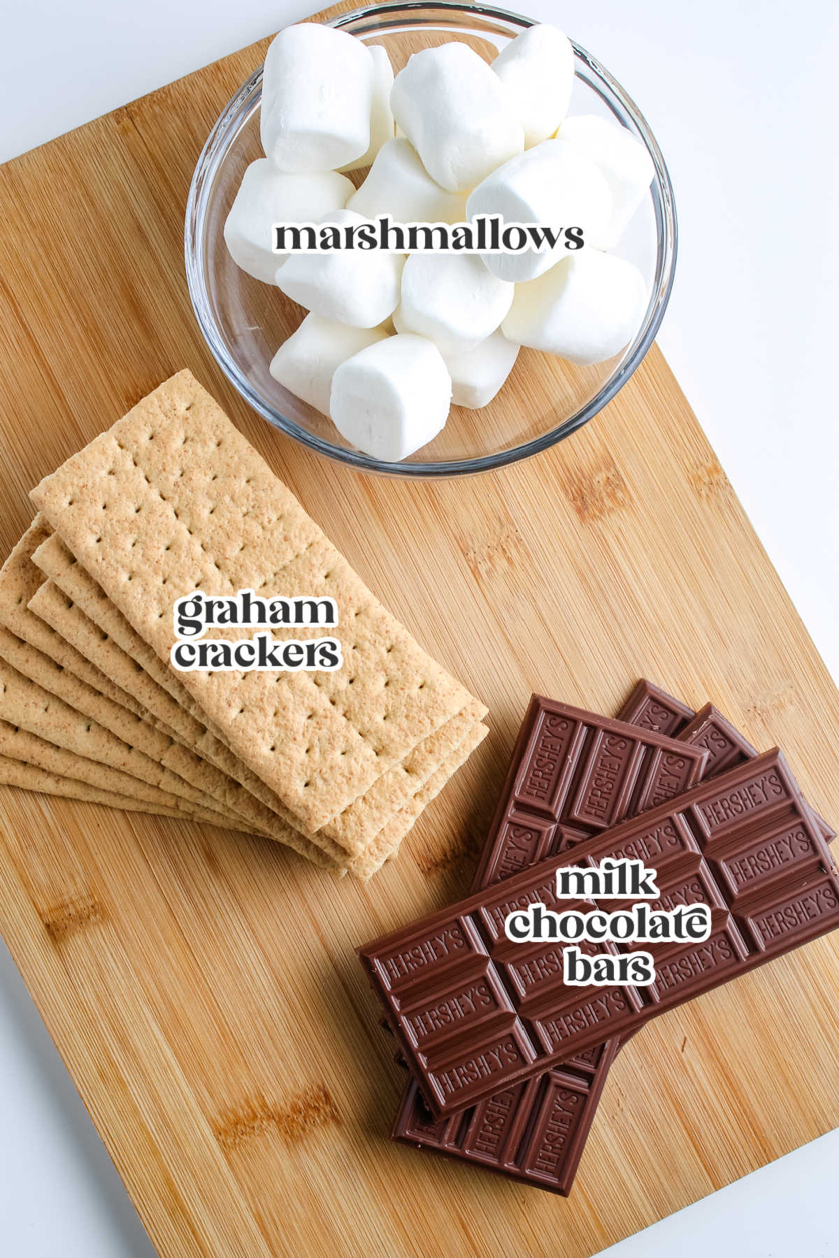 Ingredients for grilled s'mores including graham crackers, marshmallows, and milk chocolate bars on a wooden board.