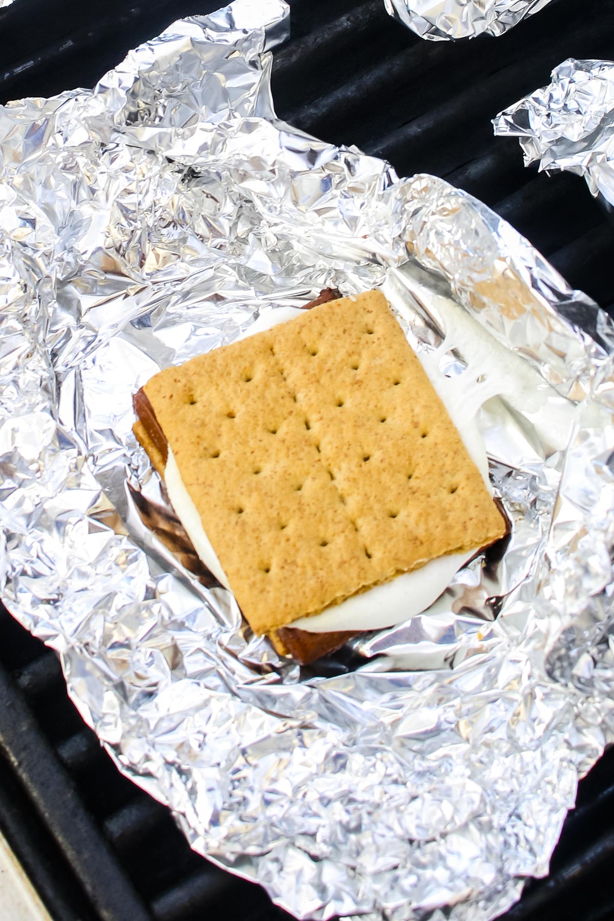 Unwrapped grilled s'more assembled with graham crackers, chocolate, and a marshmallow on aluminum foil over a grill grate.