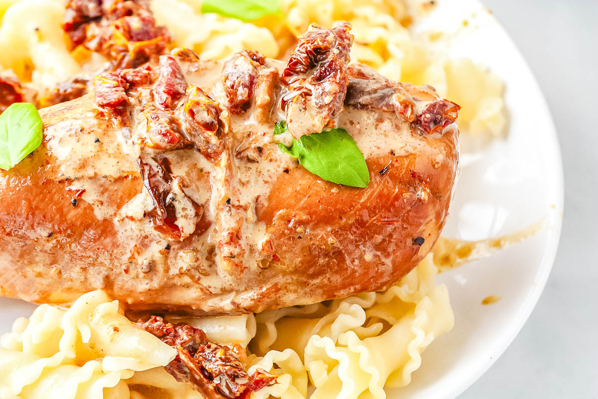 Close-up of Crockpot Marry Me Chicken topped with creamy Parmesan sauce and chopped sun-dried tomatoes over pasta.