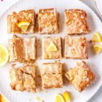 An overhead view of nine glazed lemon blondies cut into squares and arranged on a white textured serving plate with fresh lemon slices.