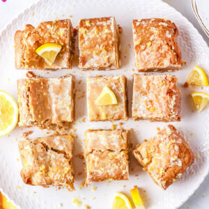 An overhead view of nine glazed lemon blondies cut into squares and arranged on a white textured serving plate with fresh lemon slices.