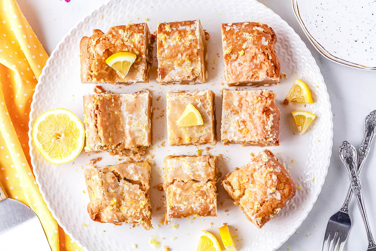 An overhead view of nine glazed lemon blondies cut into squares and arranged on a white textured serving plate with fresh lemon slices.