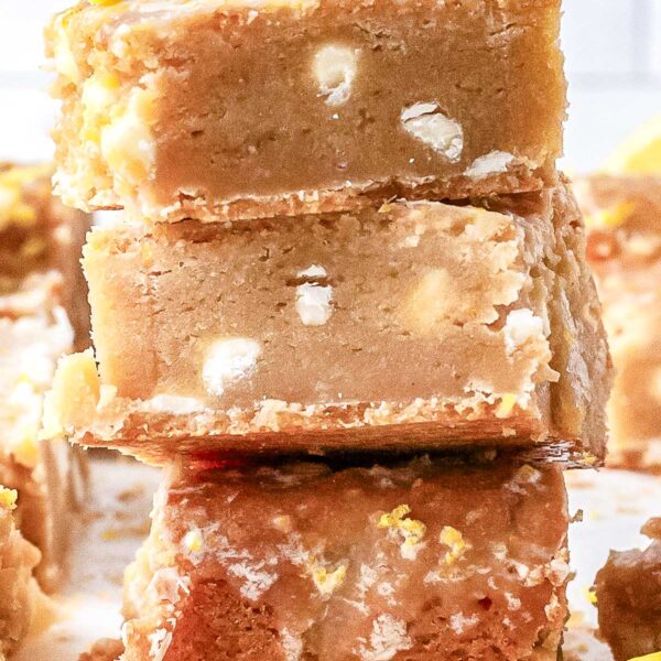 A close-up view of a stack of three baked lemon blondies showing white chocolate chips inside the chewy center, finished with a fresh lemon slice on top.
