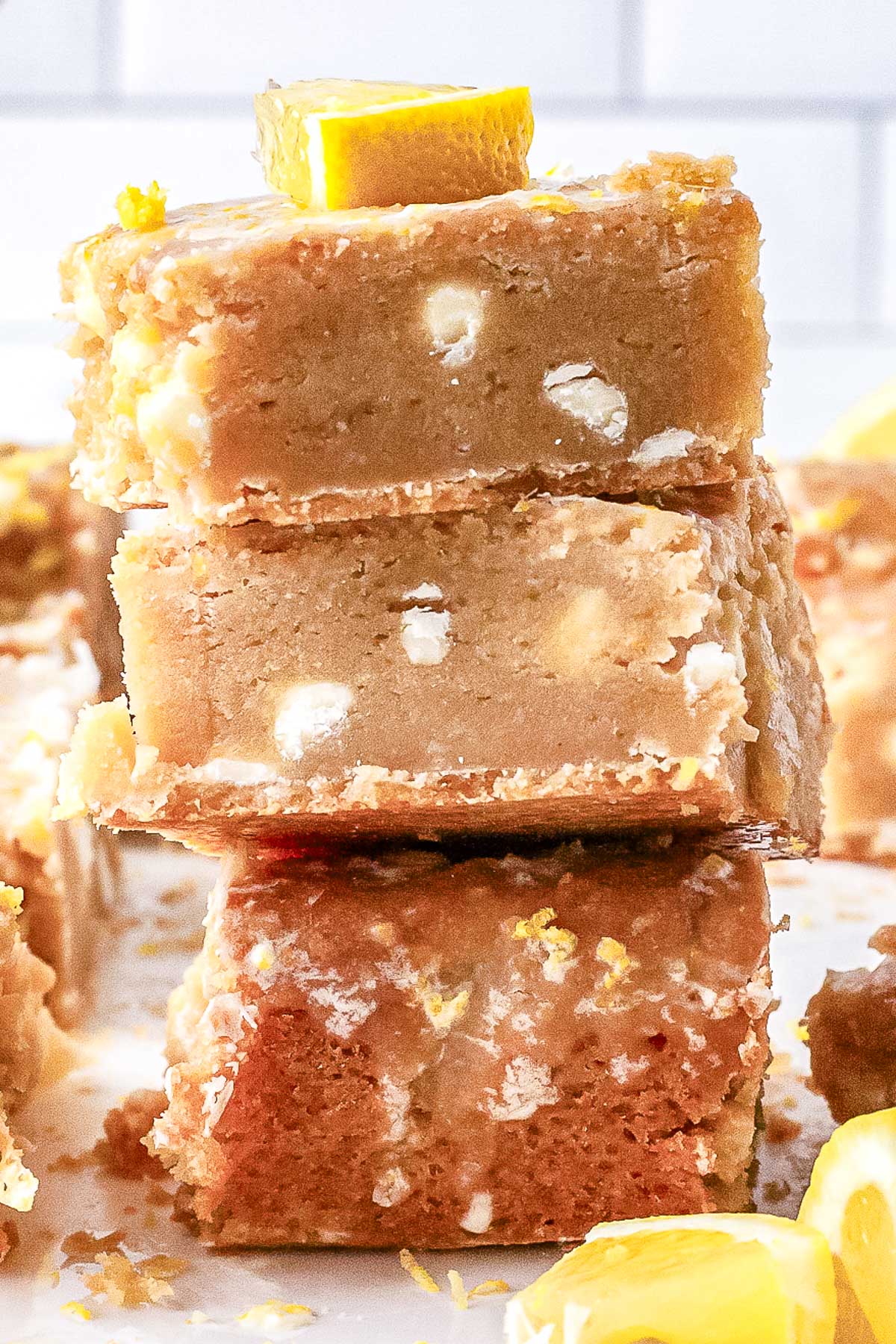 A close-up view of a stack of three baked lemon blondies showing white chocolate chips inside the chewy center, finished with a fresh lemon slice on top.