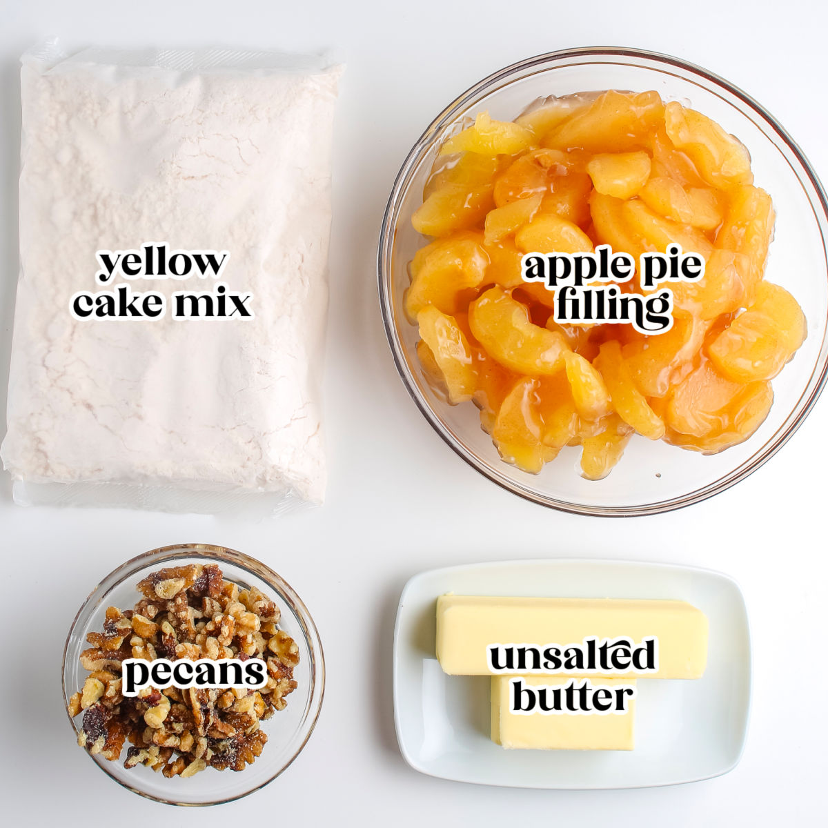 A flat lay showing labeled ingredients needed to make an apple dump cake, including a bag of yellow cake mix, a bowl of apple pie filling, chopped pecans, and unsalted butter.