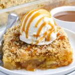 A close-up view of a square slice of warm apple dump cake topped with a scoop of vanilla ice cream and a drizzle of caramel sauce on a white plate.
